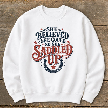 White sweatshirt with "She Believed She Could So She Saddled Up" graphic and horseshoe.