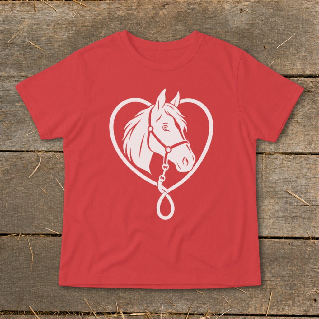 A red t-shirt lies flat on a rustic wooden surface, featuring a white graphic of a horse's head framed by a heart-shaped rope.