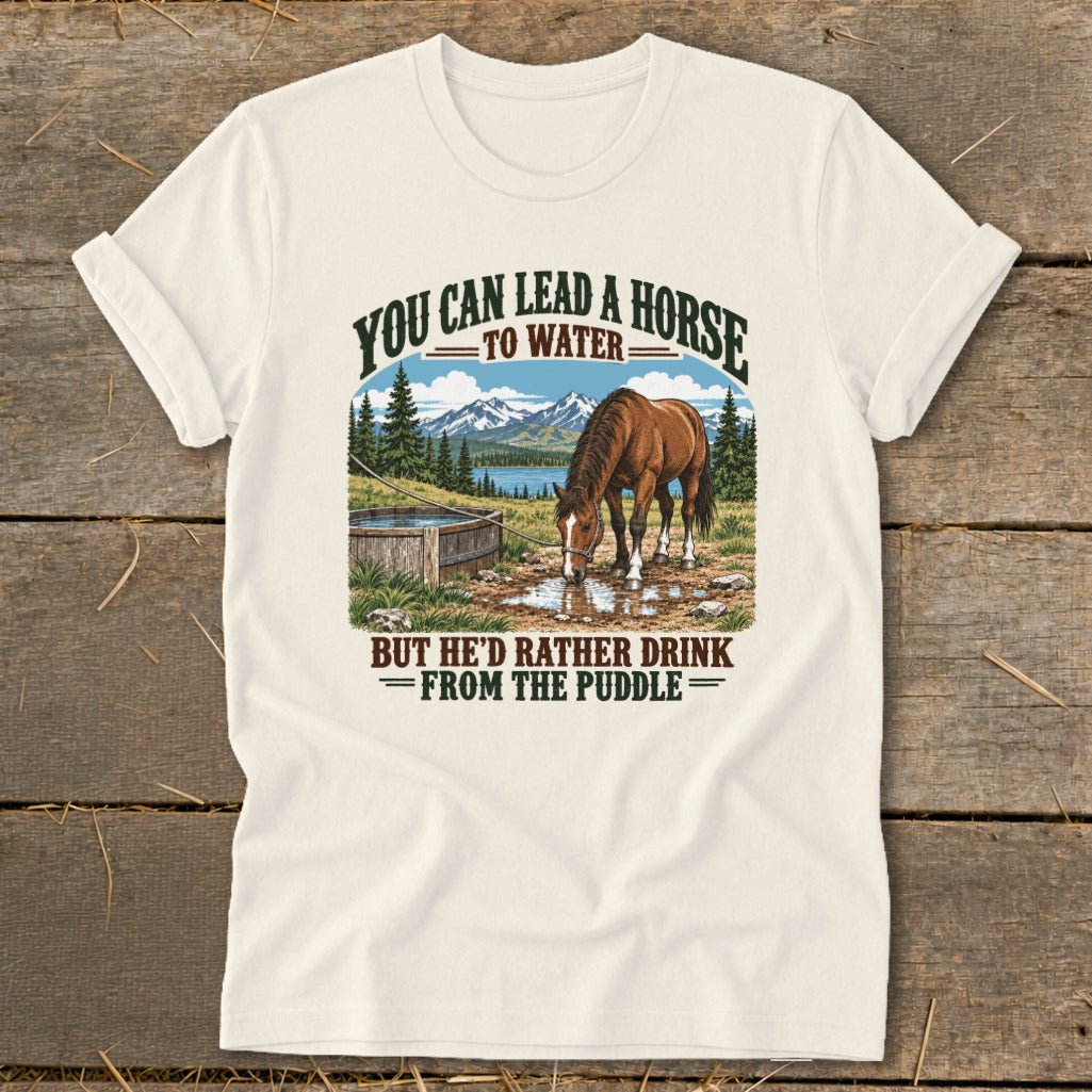 A cream-colored t-shirt featuring a retro graphic of a horse drinking from a puddle with mountains and the text You can lead a horse to water, but he'd rather drink from the puddle.
