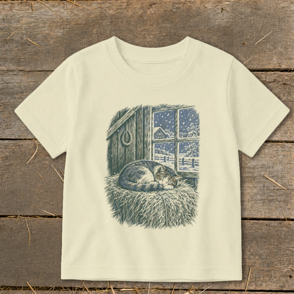 T-shirt with a barn interior and cat sleeping in hay, snow falling outside window.