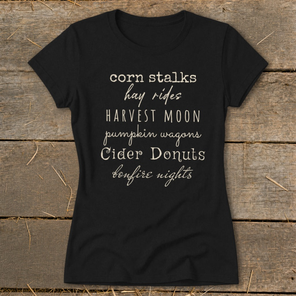 Black t-shirt with white text listing fall activities: corn stalks, hay rides, harvest moon, pumpkin wagons, cider donuts, bonfire nights.