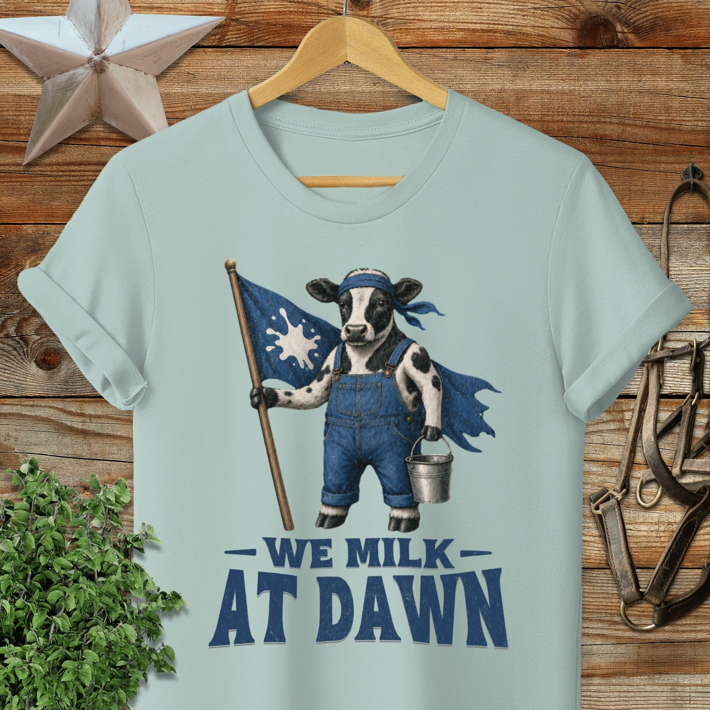 Cow wearing overalls and cape holding flag, with text "WE MILK AT DAWN".