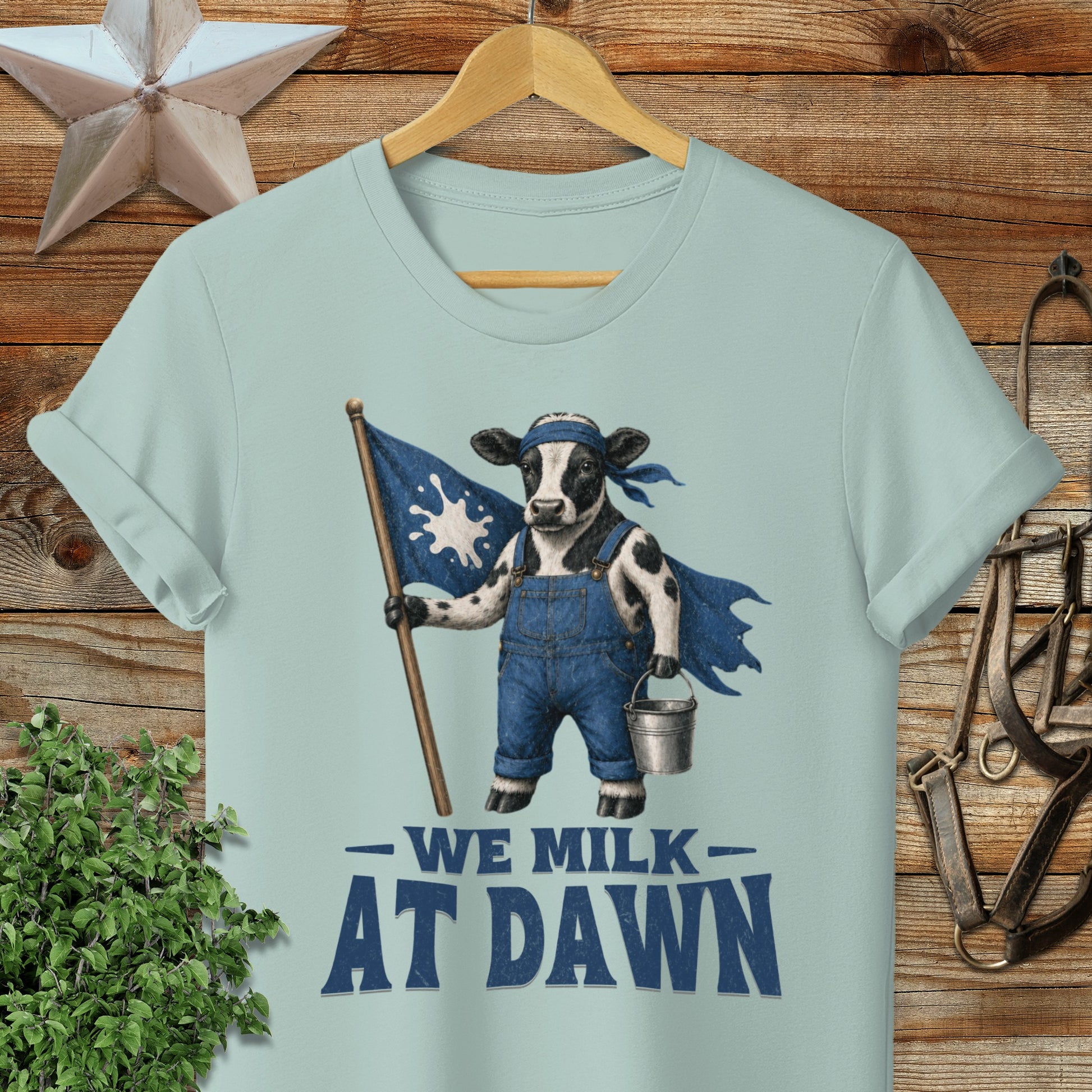Cow wearing overalls and cape holding flag, with text "WE MILK AT DAWN".