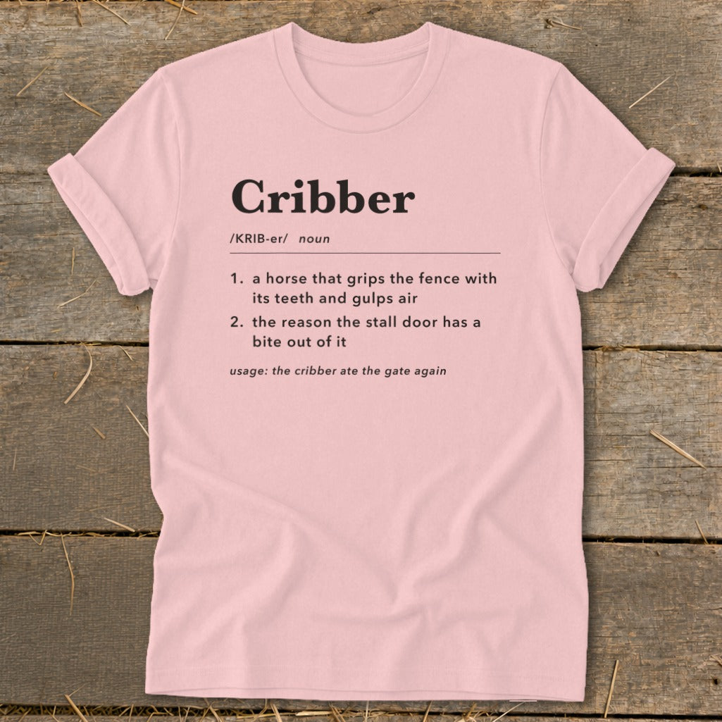 A light pink t-shirt printed with a humorous definition of the word "cribber" laid flat on a rustic wooden background.