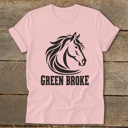 A light pink t-shirt with a black horse graphic and the text GREEN BROKE on a wooden surface.