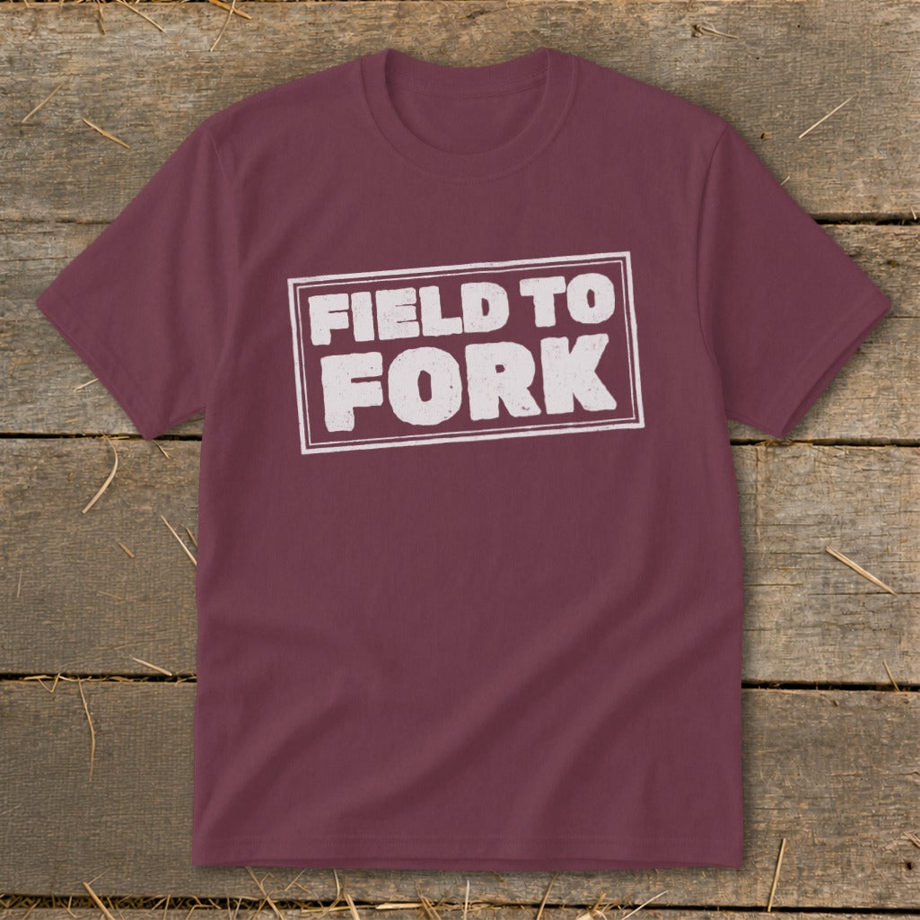 Maroon crewneck t-shirt featuring the distressed white text "Field to Fork" inside a rectangular border, displayed on a rustic wooden background.