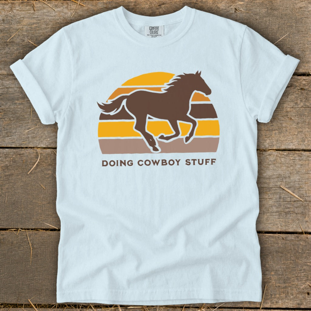 Light blue t-shirt with a brown horse silhouetted against a retro sunset and text "DOING COWBOY STUFF."