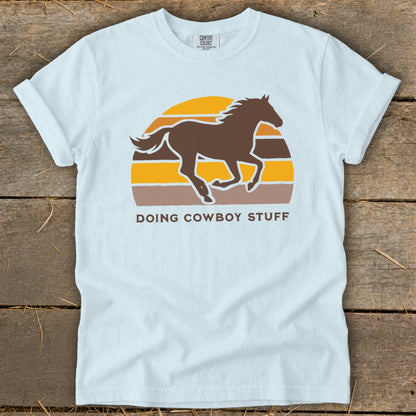 Light blue t-shirt with a brown horse silhouetted against a retro sunset and text "DOING COWBOY STUFF."
