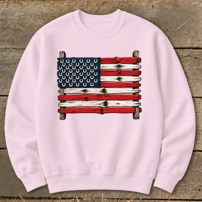 A light pink crewneck sweatshirt features a graphic of the American flag made from weathered wooden planks and horseshoes.