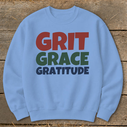 Light blue crewneck sweatshirt with colorful text reading GRIT GRACE GRATITUDE laid on wood.