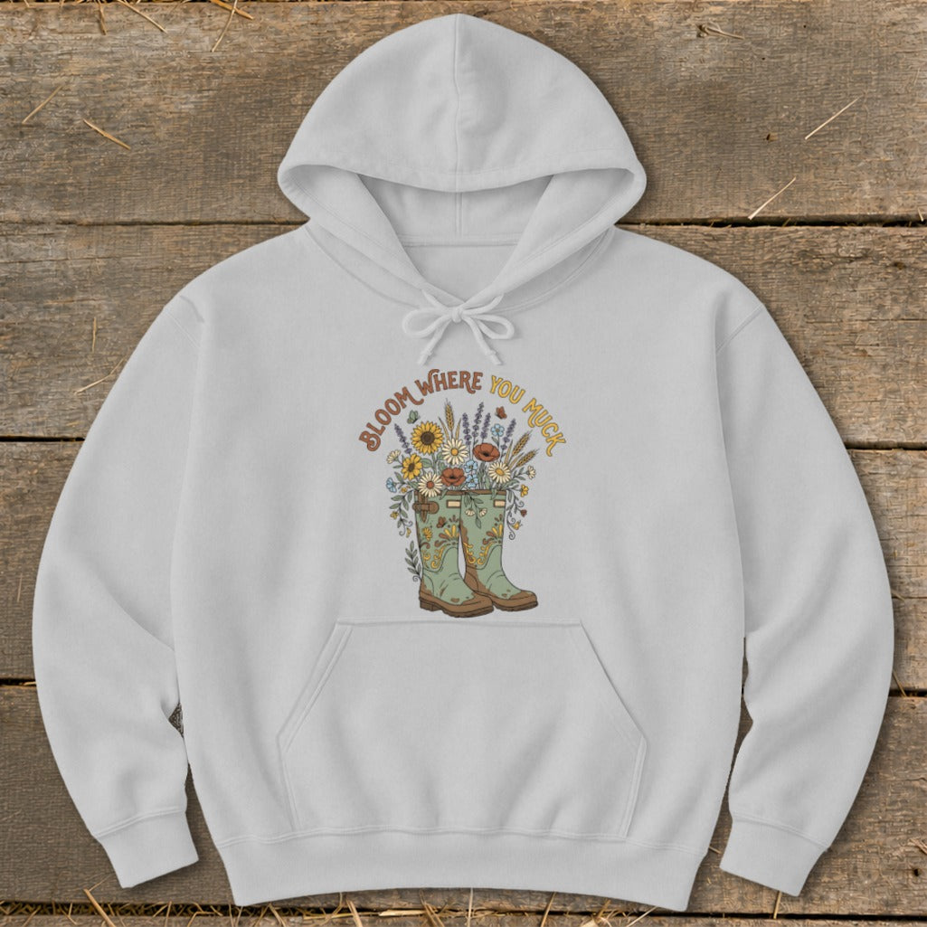 White hoodie with a graphic of boots overflowing with flowers and text "Bloom Where You Muck".