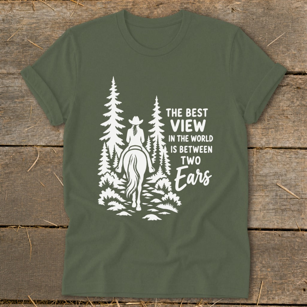 An olive green t-shirt featuring a white graphic of a person on a horse riding through a forest, alongside text reading the best view in the world is between two ears.