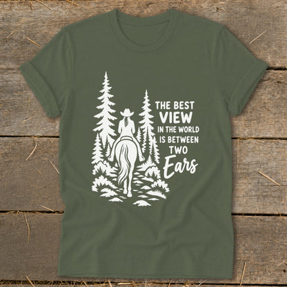 An olive green t-shirt featuring a white graphic of a person on a horse riding through a forest, alongside text reading the best view in the world is between two ears.