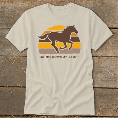 Light tan t-shirt with a brown running horse silhouette and "DOING COWBOY STUFF" text.
