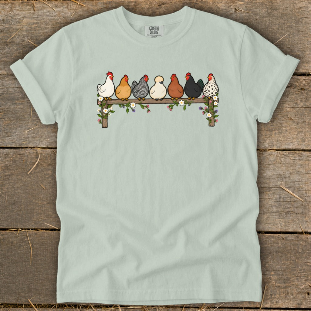 Light green crewneck t-shirt featuring an illustration of seven colorful chickens perched on a flower-covered wooden fence.