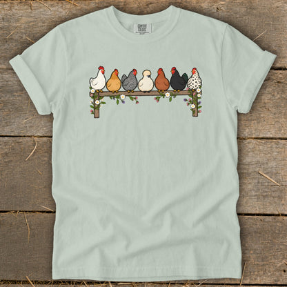 Light green crewneck t-shirt featuring an illustration of seven colorful chickens perched on a flower-covered wooden fence.