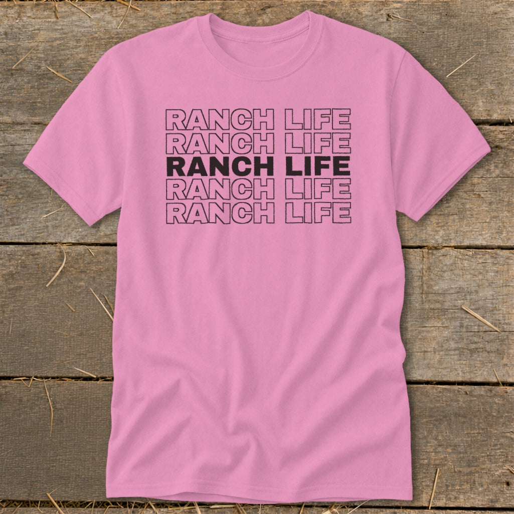 Pink short-sleeve t-shirt featuring the text Ranch Life repeated in five stacked rows, displayed on a wooden background.
