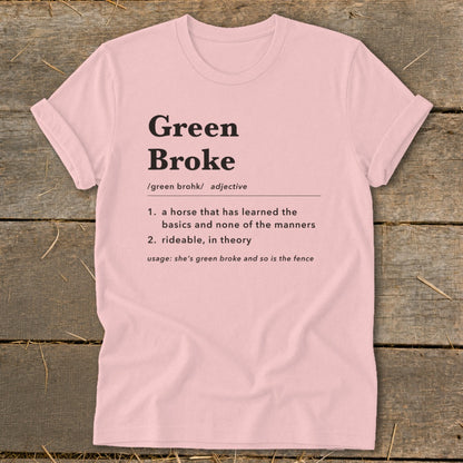 A light pink t-shirt with black text defining the term Green Broke on a rustic wooden background.