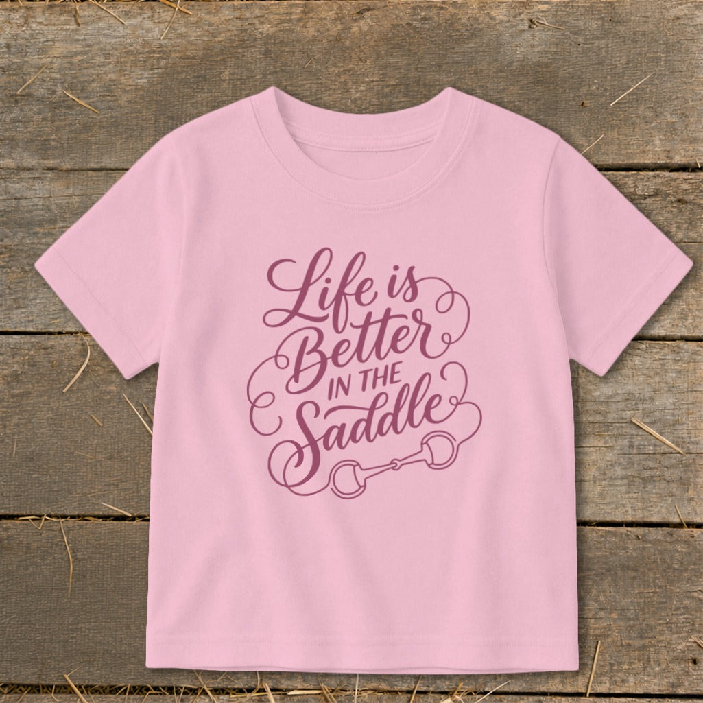 Pink t-shirt with purple text "Life is Better IN THE Saddle" and horse bit graphic.