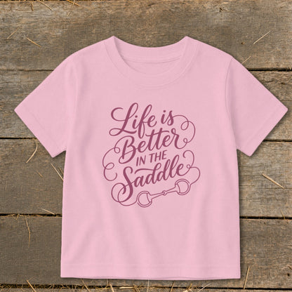 Pink t-shirt with purple text "Life is Better IN THE Saddle" and horse bit graphic.