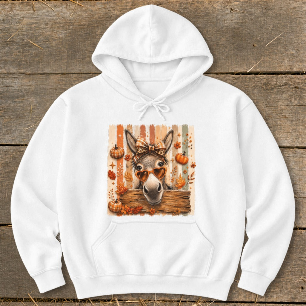 Donkey with heart sunglasses and bow behind wooden fence, surrounded by autumn leaves and pumpkins.
