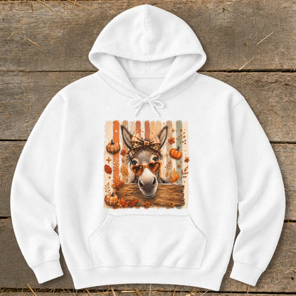 Donkey with heart sunglasses and bow behind wooden fence, surrounded by autumn leaves and pumpkins.