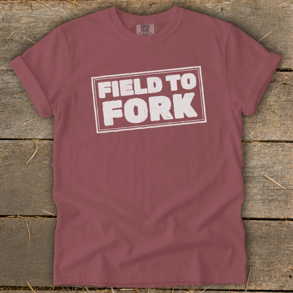 A maroon crewneck t-shirt with rolled sleeves lies flat on a rustic wooden surface, featuring the white text "FIELD TO FORK" inside a distressed rectangular box on the chest.