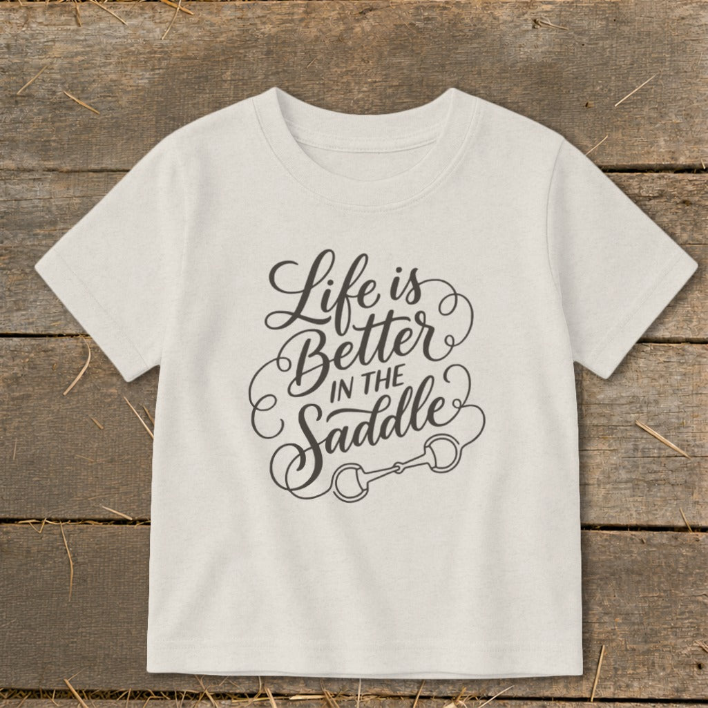 Light-colored t-shirt with "Life is Better In the Saddle" text and horse bit graphic.