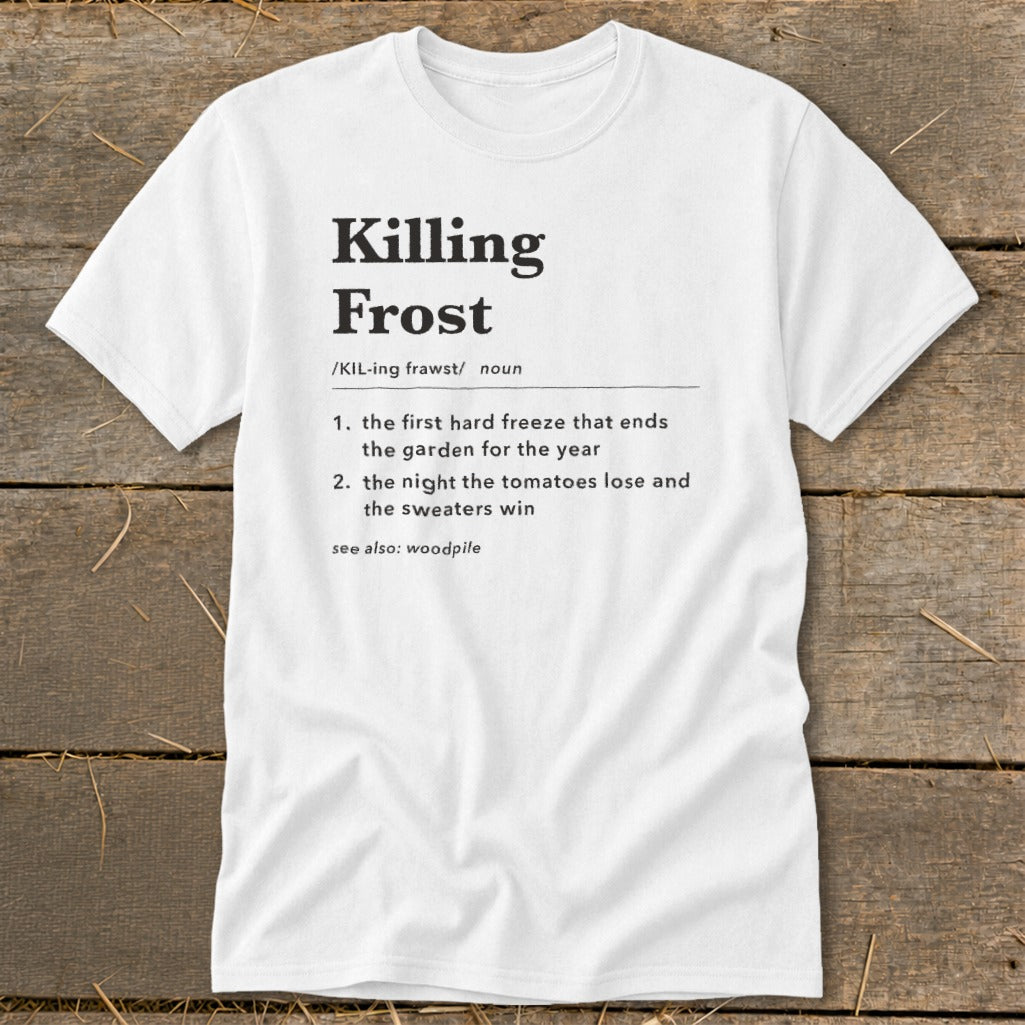 A white short-sleeved t-shirt with a humorous dictionary-style definition of Killing Frost printed in black on the chest, laid flat on a rustic wooden background.