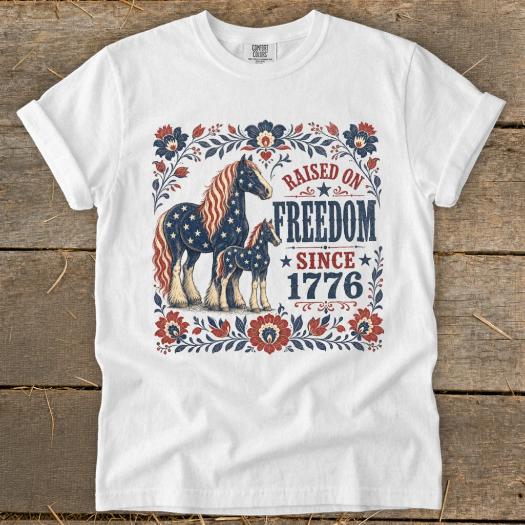 White t-shirt with patriotic horses and floral border, reads "Raised on Freedom Since 1776".
