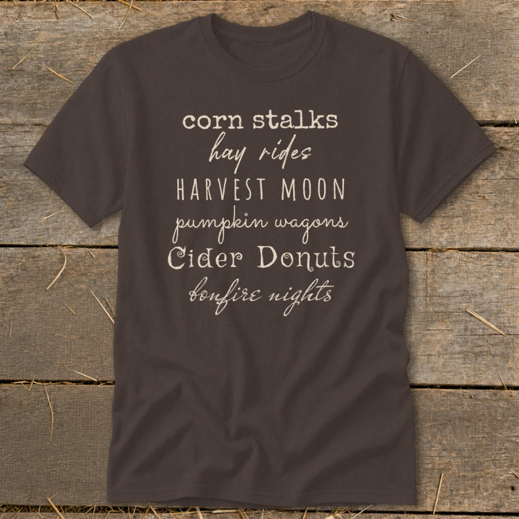 Brown t-shirt with white text listing fall activities: corn stalks, hay rides, harvest moon, pumpkin wagons, cider donuts, bonfire nights.