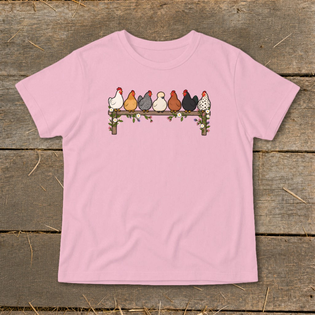 A light pink t-shirt featuring an illustration of seven colorful chickens perched on a flower-covered wooden fence.
