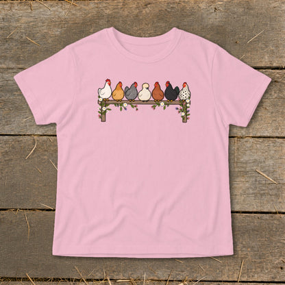 A light pink t-shirt featuring an illustration of seven colorful chickens perched on a flower-covered wooden fence.