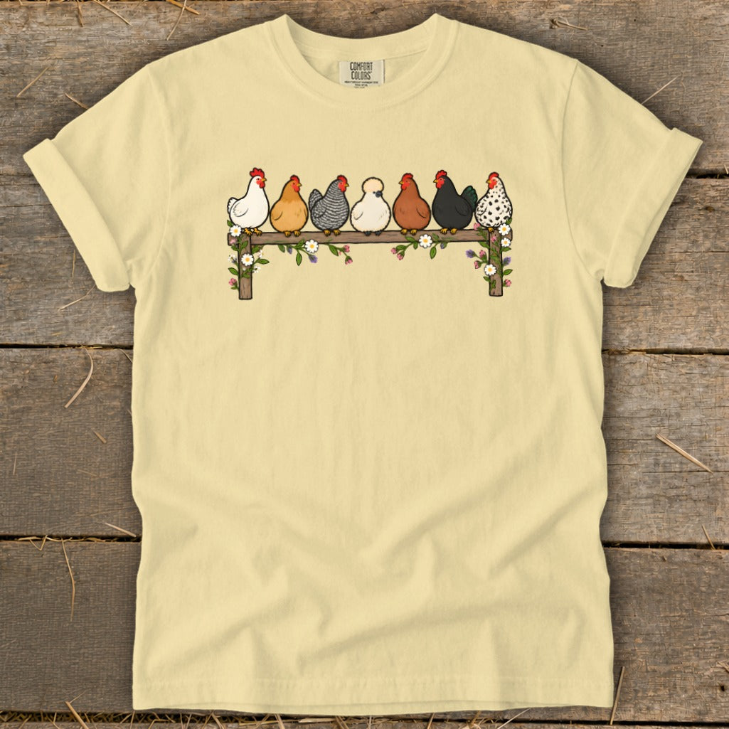 A light yellow t-shirt features a graphic of seven colorful chickens perched on a flower-covered wooden fence.