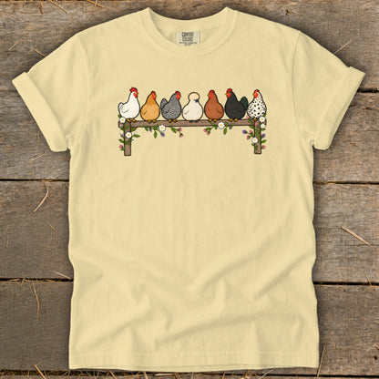 A light yellow t-shirt features a graphic of seven colorful chickens perched on a flower-covered wooden fence.