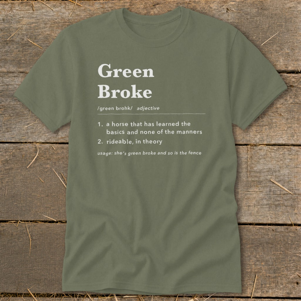An olive green t-shirt with white text defining the adjective "Green Broke" on a wooden background.