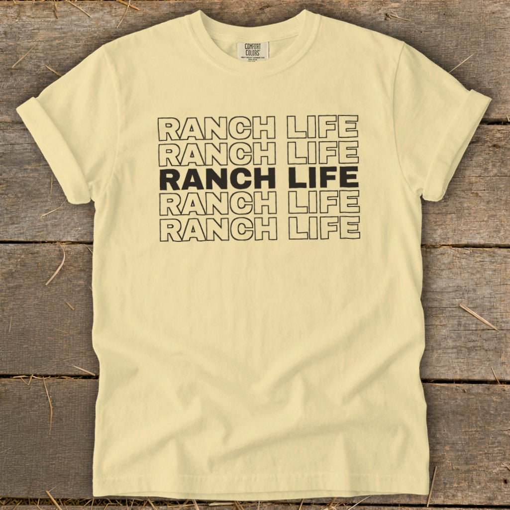 A light yellow t-shirt with rolled-up sleeves displays stacked black lettering spelling "RANCH LIFE" against a wooden background.