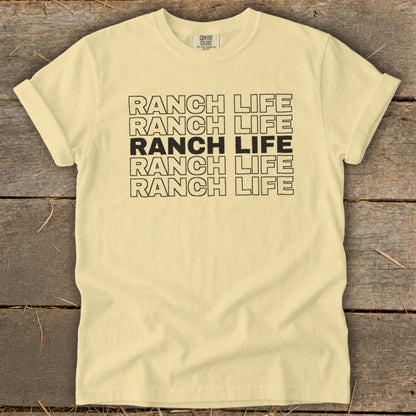 A light yellow t-shirt with rolled-up sleeves displays stacked black lettering spelling "RANCH LIFE" against a wooden background.