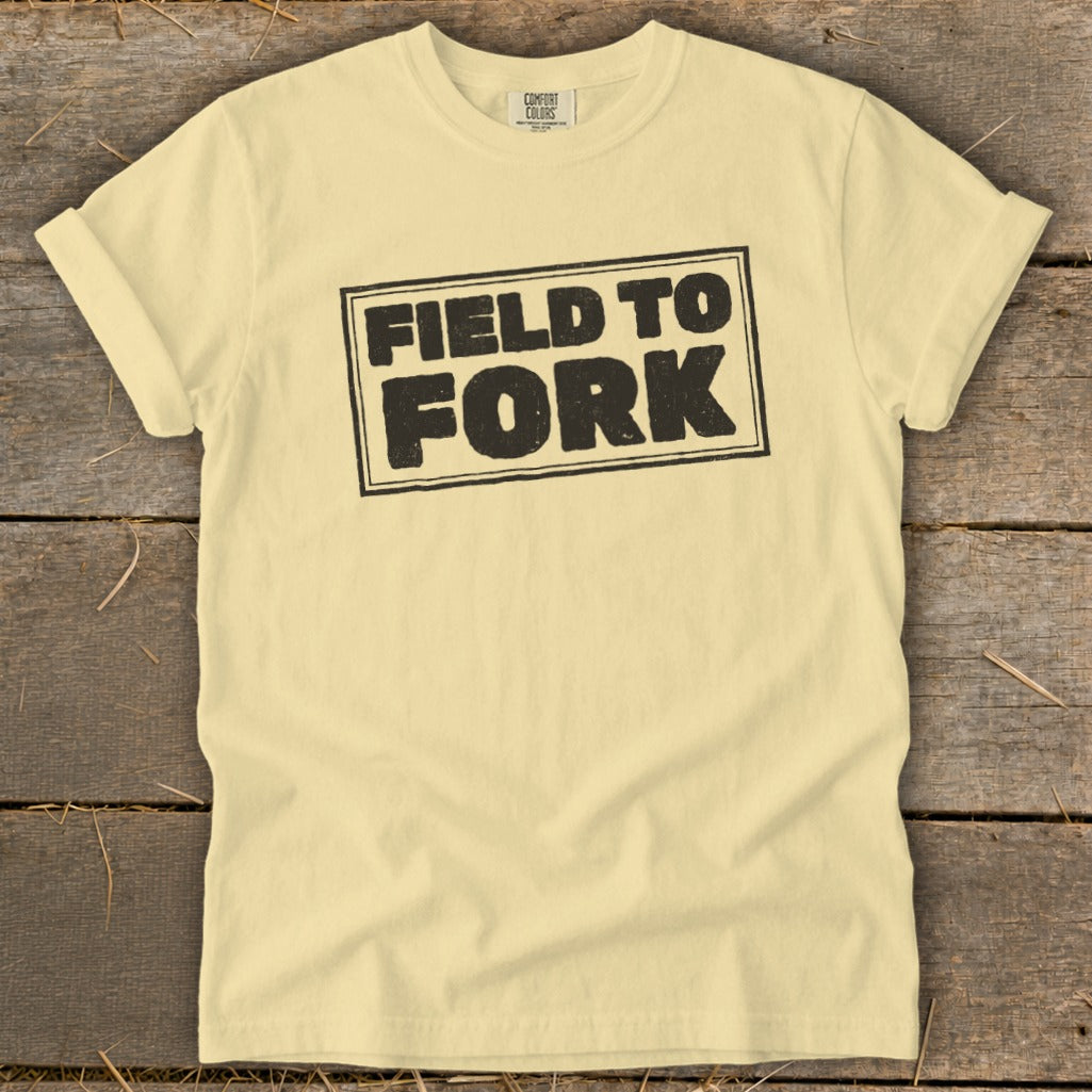 A pale yellow t-shirt laid flat on a wooden surface, featuring the black text FIELD TO FORK centered on the chest.