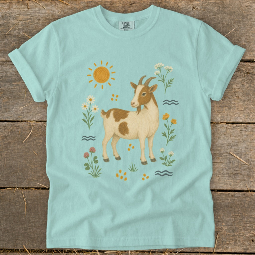 Light blue t-shirt with a tan and white goat surrounded by flowers, sun, and waves.