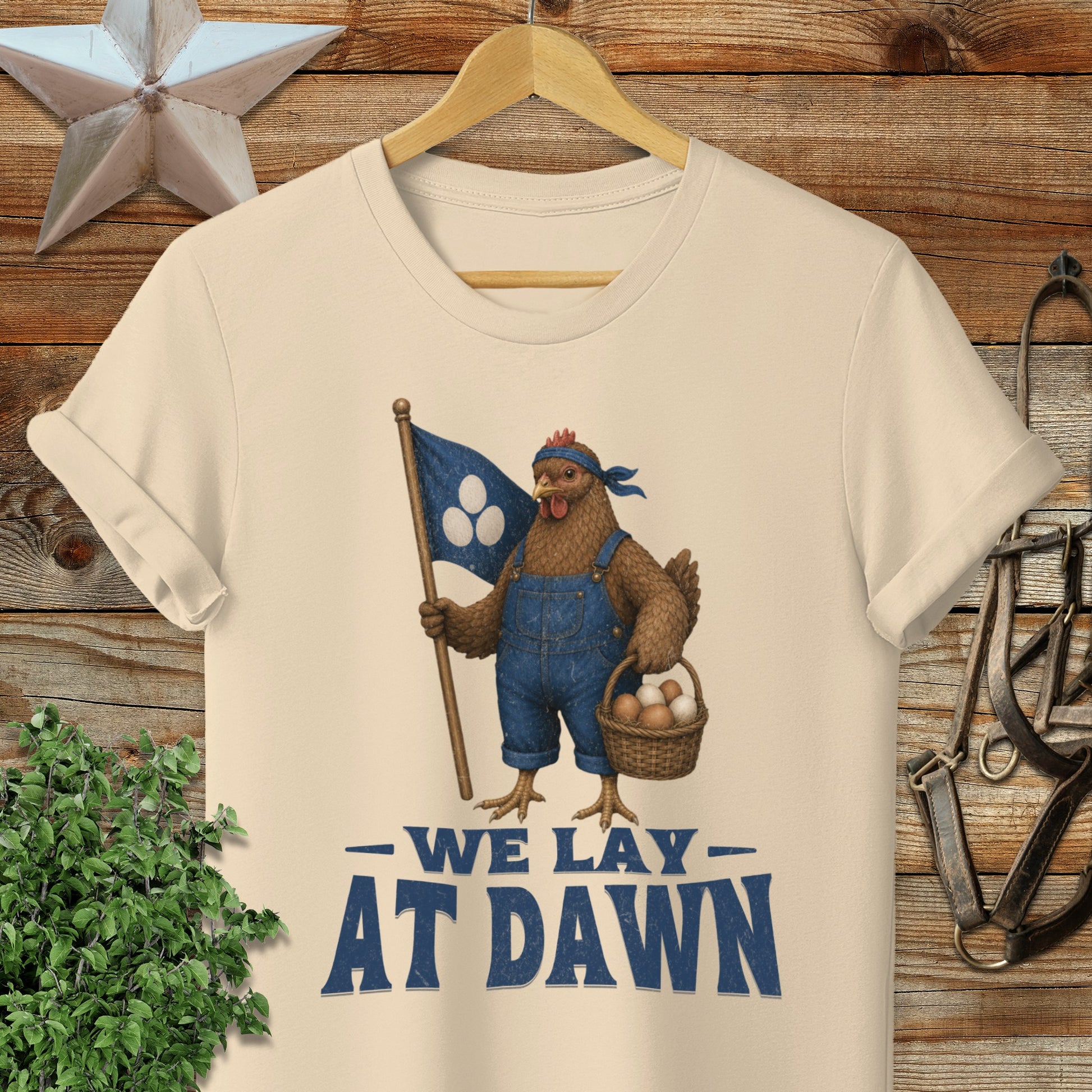 Cream t-shirt with chicken in overalls holding flag and basket, reads "We Lay At Dawn".