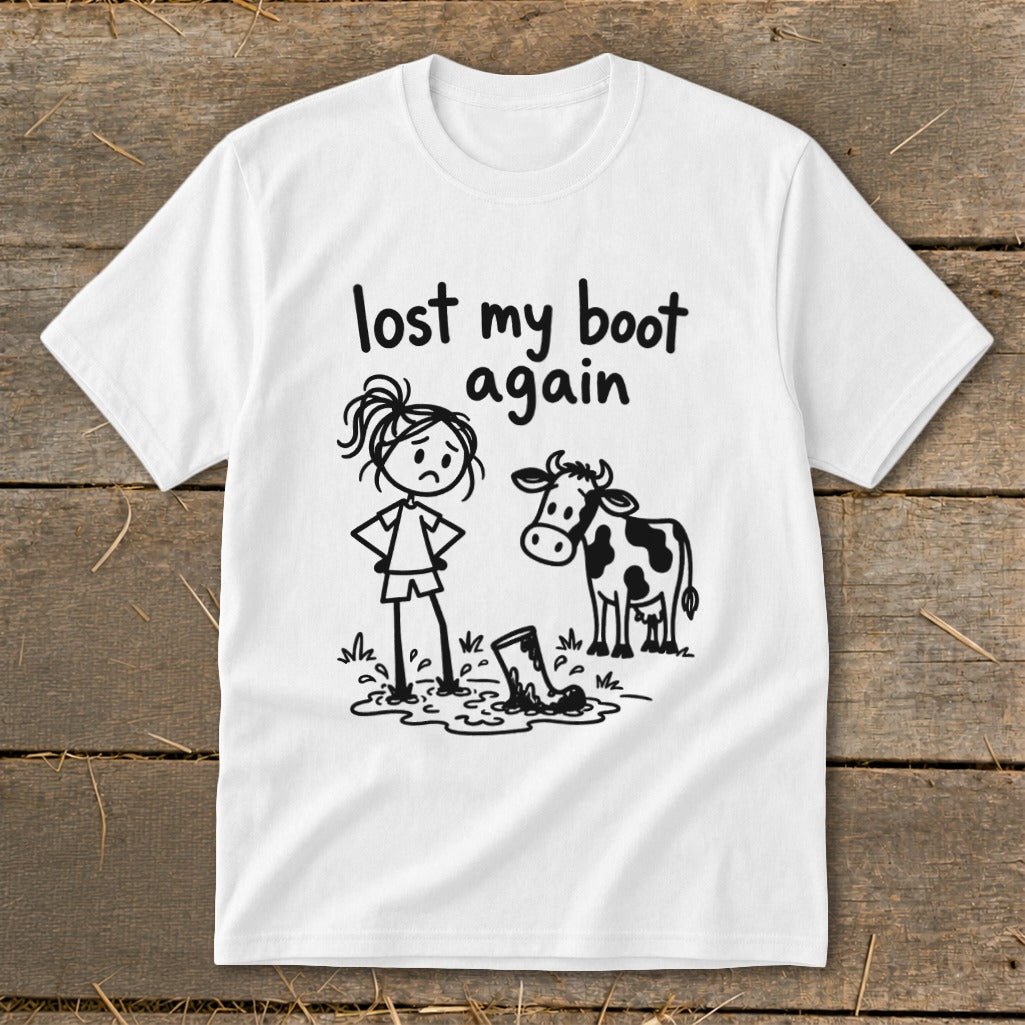 A white short-sleeved t-shirt features a cartoon graphic of a sad stick-figure girl, a cow, and text that reads lost my boot again.