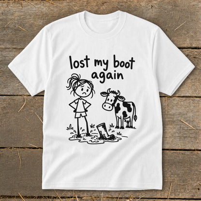 A white short-sleeved t-shirt features a cartoon graphic of a sad stick-figure girl, a cow, and text that reads lost my boot again.