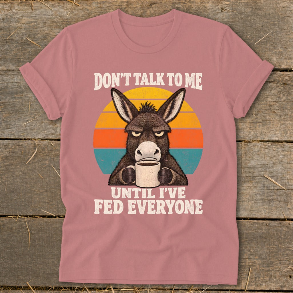 Dusty pink t-shirt featuring a graphic of an annoyed donkey holding a mug, with text saying "DON'T TALK TO ME UNTIL I'VE FED EVERYONE."