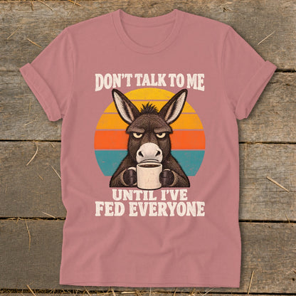 Dusty pink t-shirt featuring a graphic of an annoyed donkey holding a mug, with text saying "DON'T TALK TO ME UNTIL I'VE FED EVERYONE."