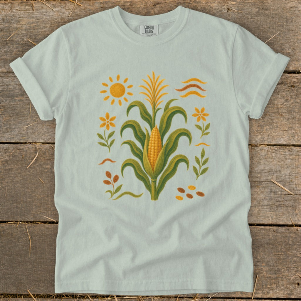 Light blue t-shirt with a corn stalk, sun, flowers, and seeds graphic.