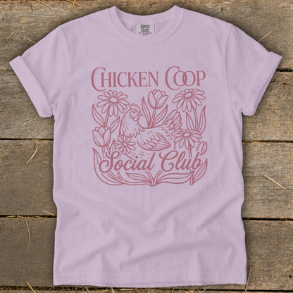 Lavender t-shirt with a pink line drawing of a chicken among flowers and text "CHICKEN COOP Social Club".