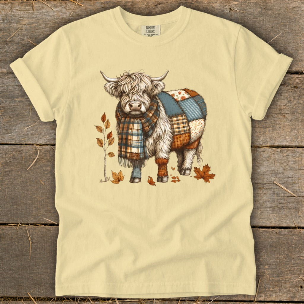 Cream t-shirt with a Highland cow illustration wearing a scarf and patchwork blanket.