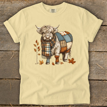 Cream t-shirt with a Highland cow illustration wearing a scarf and patchwork blanket.