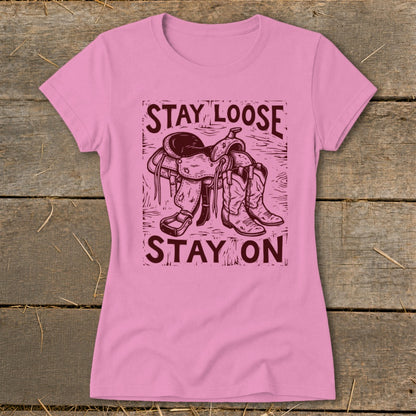 Pink t-shirt with a woodcut-style graphic of a saddle and boots reading "STAY LOOSE STAY ON".